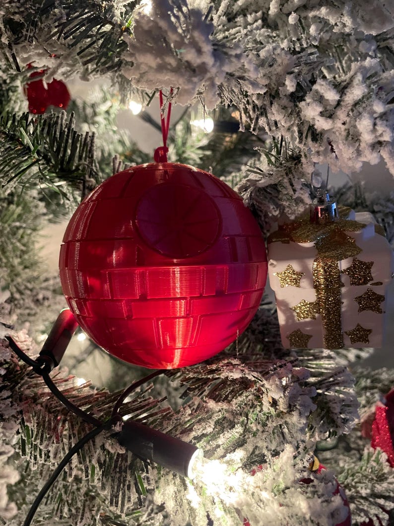 Pode incluir: Um enfeite de Natal vermelho em forma de Estrela da Morte pendurado numa &aacute;rvore de Natal nevada. O enfeite &eacute; uma esfera com detalhes na superf&iacute;cie. Um enfeite branco em forma de presente com detalhes dourados tamb&eacute;m &eacute; vis&iacute;vel.