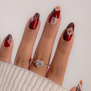 May include: Almond-shaped fingernails featuring a patchwork design in shades of deep red, dark brown, and cream, accented with black stitch-like lines and small leaf motifs. A gold-toned ring with a heart-shaped clear stone sits on the finger.