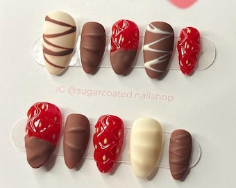 May include: A set of ten artificial nails with a dessert theme. Designs include chocolate-covered strawberries, chocolate bars, and white chocolate with brown stripes. The nails are displayed on a white card with the text "sugarcoated" and an Instagram handle.