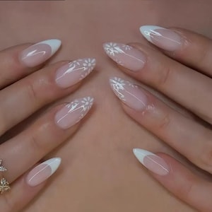 May include: Close-up of a set of manicured nails. The nails are oval-shaped with a clear base, white tips, and delicate white daisy designs. The nails are on a light skin tone background.