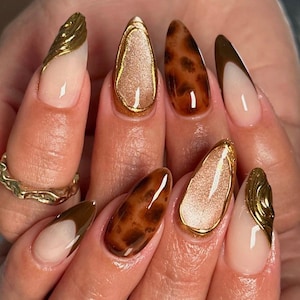 May include: Almond-shaped nails featuring a variety of designs including brown leopard print, solid brown French tips, textured gold metallic accents, and shimmering champagne-coloured centres outlined in gold, paired with a textured gold ring.