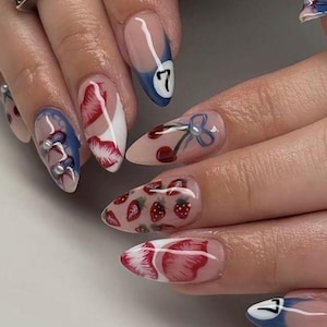 May include: A close-up of a hand with long, almond-shaped nails. The nails feature various designs, including strawberries, cherries, and a blue and white design with the number 7. Some nails have pearl accents.