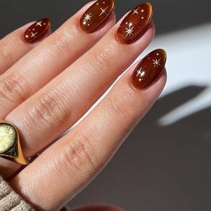 May include: Oval-shaped fingernails featuring a glossy, deep brown cat-eye polish finish. Each nail is decorated with small, white, hand-painted starburst designs and tiny dots, creating a celestial pattern across the dark, shimmering brown surface.