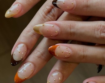 May include: Almond-shaped nails featuring a clear base coat with multi-colored French tips in shades of brown, tan, and orange. Each nail is decorated with small, metallic gold studs placed sporadically across the clear nail bed surface.