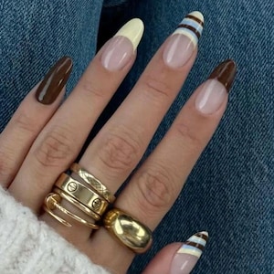 May include: Close-up of a hand with long, almond-shaped nails. Some nails are painted solid brown, others have a light yellow or brown tip, and some feature blue and white stripes. Several gold rings adorn the fingers.