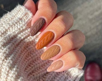 May include: Almond-shaped fingernails featuring a gradient of brown tones, ranging from dark chocolate to light beige. Each nail displays a raised, textured cable knit pattern that mimics the appearance of a sweater fabric.
