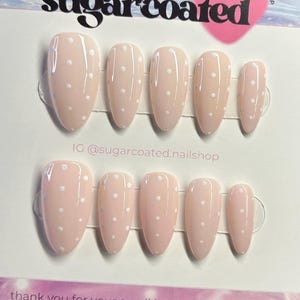 May include: A set of ten almond-shaped press-on nails in a pale pink shade, each decorated with small white polka dots. The nails are displayed on a white card with the text "sugarcoated" and an Instagram handle.