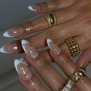 May include: Almond-shaped nails featuring a white French tip and a nude base, decorated with small silver starburst accents and tiny rhinestones. Three gold-toned rings are worn on the fingers, including a smooth band, a beaded band, and a square-cut stone band.