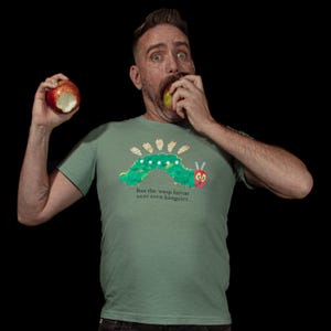 May include: A light green t-shirt featuring a cartoon caterpillar design with the text "But the wasp larvae were even hungrier." The person in the image is eating an apple.
