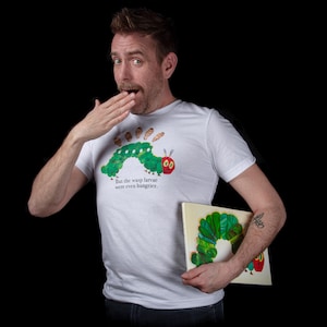 以下が含まれることがあります： 緑色の毛虫のグラフィックと「しかし、スズメバチの幼虫はさらに空腹でした。」というテキストが書かれた白いTシャツ。人がそのTシャツを着て、同じような毛虫のグラフィックが描かれた本を持っています。