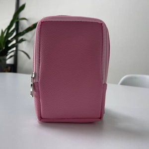 May include: A pink zippered pouch with a textured finish. The pouch is made of a soft, faux leather material.