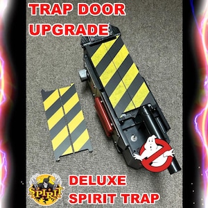 May include: Deluxe Spirit Trap with a black and yellow striped door upgrade. The trap features a black body with red and silver accents, and a Ghostbusters logo. The text "TRAP DOOR UPGRADE" and "DELUXE SPIRIT TRAP" are visible.