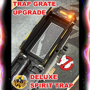 May include: A deluxe Spirit Trap upgrade with the text "TRAP GRATE UPGRADE" in red. The trap is black and yellow with a silver grate and an illuminated orange light. The Ghostbusters logo is visible.