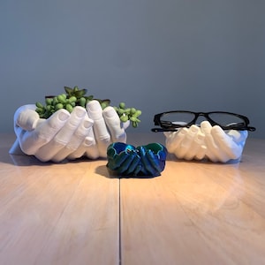 May include: Two white ceramic hand-shaped planters, one holding a small succulent plant, the other holding a pair of black eyeglasses. A small blue and green decorative object sits in front of the planters.