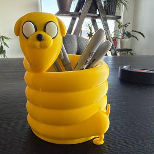May include: A bright yellow cylindrical pencil holder shaped like a coiled cartoon dog with a rounded head, large white eyes, and a small black nose. The body forms a hollow container for pens and markers, featuring a small tail at the base.