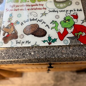 May include: A white cutting board featuring a green Grinch character and a message that reads "Dear Mr. Grinch I gift you with this goodie tray So you don't wreck my Christmas day. Something warm for you to drink A treat baked just for you Treat for Max"