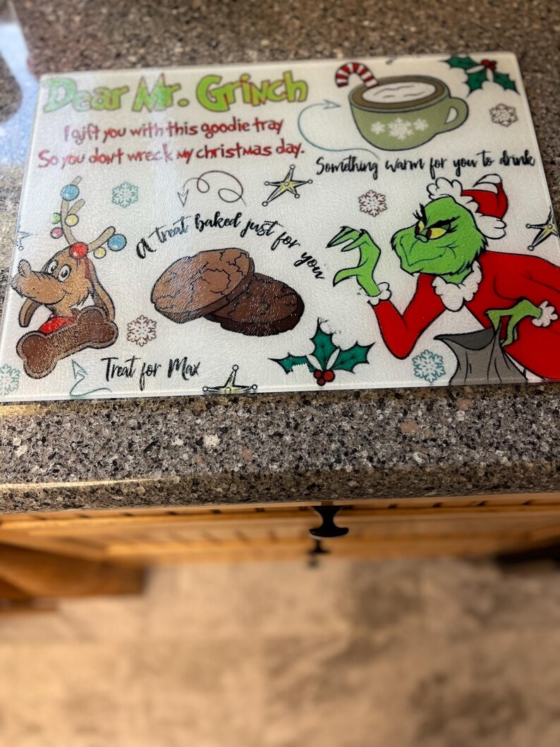 May include: A white cutting board featuring a green Grinch character and a message that reads "Dear Mr. Grinch I gift you with this goodie tray So you don't wreck my Christmas day. Something warm for you to drink A treat baked just for you Treat for Max"