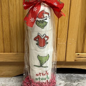 May include: Three rolls of toilet paper wrapped in clear plastic with a red and white snowflake ribbon. The toilet paper has a Grinch Christmas theme with images of the Grinch wearing a Santa hat and a red and white suit. The bottom of the package is decorated with pink tissue paper. The text "Stink Stank" is printed on the toilet paper.