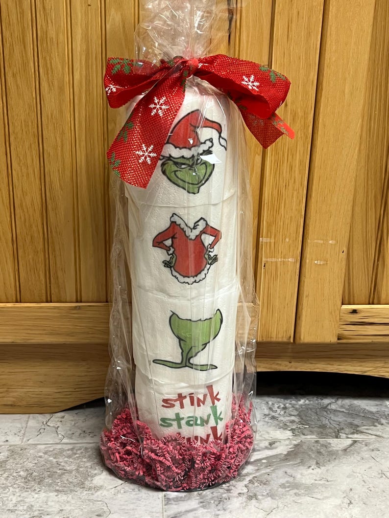 May include: Three rolls of toilet paper wrapped in clear plastic with a red and white snowflake ribbon. The toilet paper has a Grinch Christmas theme with images of the Grinch wearing a Santa hat and a red and white suit. The bottom of the package is decorated with pink tissue paper. The text "Stink Stank" is printed on the toilet paper.