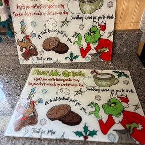 May include: Two white ceramic chopping boards with a Grinch theme. The boards feature a green Grinch character, a reindeer, and a treat baked just for you. The text on the boards reads "Dear Mr. Grinch I gift you with this goodie tray So you don't wreck my Christmas day. Something warm for you to drink A treat baked just for you Treat for Max".