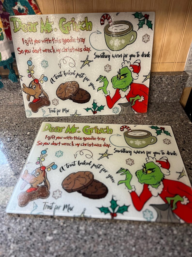 May include: Two white ceramic chopping boards with a Grinch theme. The boards feature a green Grinch character, a reindeer, and a treat baked just for you. The text on the boards reads "Dear Mr. Grinch I gift you with this goodie tray So you don't wreck my Christmas day. Something warm for you to drink A treat baked just for you Treat for Max".