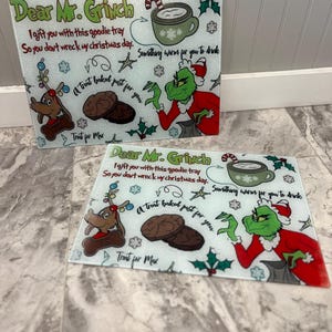 May include: Two glass chopping boards with a Grinch theme. The boards feature a green Grinch character, a reindeer, and a Christmas tree. The text on the boards reads "Dear Mr. Grinch I gift you with this goodie tray So you don't wreck my Christmas day. A treat baked just for you Treat for Max Something warm for you to drink".