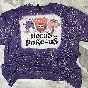 Pode incluir: Camiseta roxa com um gráfico branco com personagens de desenhos animados e o texto "Hocus Poke-us". A camisa tem um design desbotado e salpicado.