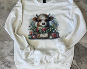 May include: White Christmas sweatshirt with a cute cartoon cow wearing a red bow and a wreath of lights. The cow is surrounded by presents and the text "Merry Christmas" is printed on a sign.