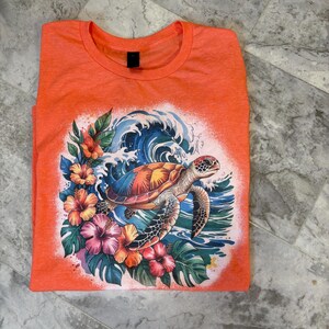 Könnte beinhalten: Korallenfarbenes T-Shirt mit einem farbenfrohen Aufdruck einer Meeresschildkröte in Ozeanwellen, umgeben von tropischen Blumen und Blättern. Das Design umfasst Blau-, Orange-, Rosa- und Grüntöne und erzeugt eine sommerliche Atmosphäre.