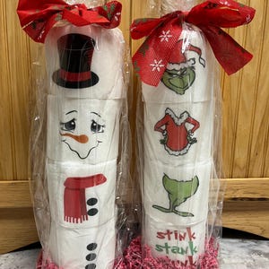 May include: Two rolls of toilet paper wrapped in clear plastic and decorated with Christmas themes. One roll is decorated with a snowman and the other with the Grinch. Both rolls are wrapped with red ribbon and have pink tissue paper at the bottom.