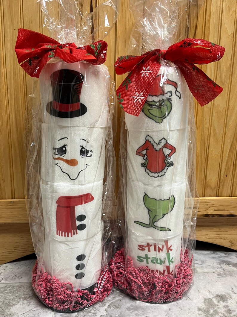 May include: Two rolls of toilet paper wrapped in clear plastic and decorated with Christmas themes. One roll is decorated with a snowman and the other with the Grinch. Both rolls are wrapped with red ribbon and have pink tissue paper at the bottom.