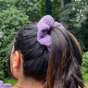 May include: A lilac crochet scrunchie secures a ponytail. The scrunchie is made of a textured yarn and matches the straps of a crochet top. The background features lush green foliage.