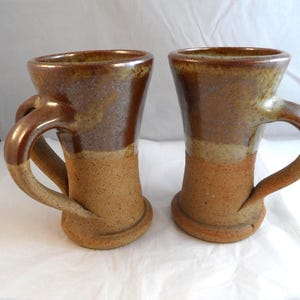 May include: Two tall ceramic mugs featuring a two-tone glaze. The upper portion of each mug has a glossy, dark brown finish, while the lower section and base exhibit a textured, speckled light tan clay surface with a flared bottom rim.