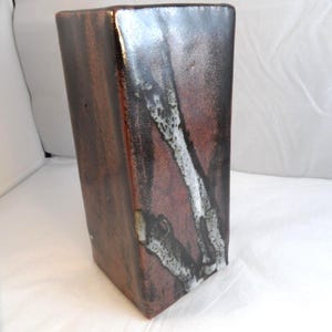 May include: A rectangular ceramic vase with a dark brown and reddish-brown glaze. The vase has a white and black abstract design on one side. The vase is approximately 25 cm tall.