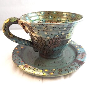 May include: A handmade ceramic teacup and saucer set. The cup and saucer are teal with a speckled pattern and leaf designs. The handle is green and yellow. The set is designed for tea or coffee.