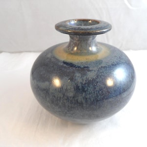 May include: A round ceramic vase with a narrow neck and a wide, flat rim. The surface features a mottled glaze with deep blue, grey, and charcoal tones, transitioning into a soft, warm tan band around the base of the neck with a glossy finish.