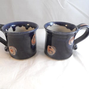 May include: Two ceramic mugs with a deep navy blue glaze and a white interior. The exterior features a smooth finish with scattered, circular orange and tan accents. Each mug has a sturdy, curved handle and a wide, flared rim design.