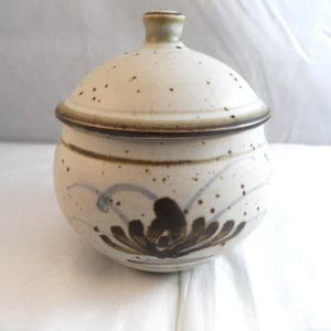 May include: A round ceramic lidded jar with a speckled off-white glaze. The body features a hand-painted dark brown floral motif and faint blue curved lines. The lid has a small knob handle and a dark brown rim, matching the jar's upper edge.