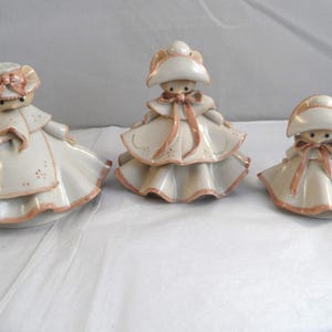 May include: Three ceramic figurines of girls in white dresses and hats with peach-coloured accents. The figurines have a vintage aesthetic, with detailed facial features and bows. The dresses have a layered design, and the hats have a rounded shape.