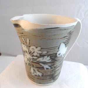 May include: A ceramic jug with a white interior and handle. The exterior has a textured, grey-brown design with white floral and butterfly accents. The jug features a pouring spout.