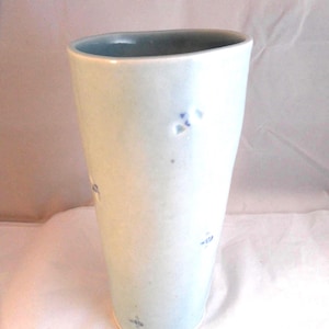 May include: A tall, pale blue ceramic vase with a slightly flared top. The vase has a smooth surface with subtle blue markings and a darker blue interior. The vase is approximately 25 cm tall, suitable for displaying flowers or as a decorative item.