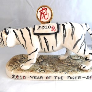 May include: A ceramic white tiger figurine with black stripes standing on a textured base. A saddle on its back features the text 2010 and a red Chinese character, topped with a circular gold-rimmed medallion. The base reads 2010-YEAR OF THE TIGER-2010.
