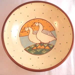 May include: A round ceramic bowl with a cream-coloured interior featuring a central illustration of two white geese standing on a green patch of grass against a yellow and blue background. The bowl has a brown rim and small green polka dots.