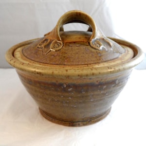 May include: A round, lidded ceramic bowl featuring a speckled, earthy brown and tan glaze. The lid includes a prominent, arched handle and subtle, embossed leaf-like decorative details. The bowl has a wide, tapered body with a smooth rim.