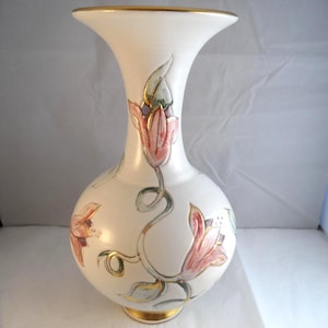 May include: A white ceramic vase with a flared rim and a rounded body, featuring hand-painted pink and green floral motifs with gold-painted accents on the flowers, leaves, and the top and bottom edges of the vessel.