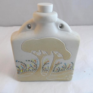 May include: A ceramic bottle with a light beige finish and a white stopper. The bottle features a stylized tree design in a lighter beige colour, with colourful dots at the base of the tree. The bottle has a square shape.