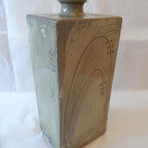 May include: A tall, square-shaped ceramic bottle with a muted olive-green glaze. The exterior features carved, sweeping arch patterns and small, repeating leaf-like impressions on each side. It has a short, narrow neck and a rounded rim.