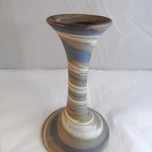 May include: A ceramic candlestick holder featuring a marbled pattern of blue, green, white, and brown tones. The object has a flared top opening, a slender cylindrical neck, and a wide, circular base, all finished with a smooth, matte texture.
