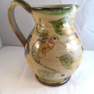 May include: A ceramic pitcher with a cream-coloured base featuring hand-painted illustrations of a hummingbird, green leaves, and yellow flowers. The pitcher has a curved handle, a wide body, and a dark brown rim at the base and top edge.