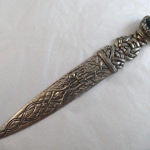 May include: A silver-toned metal letter opener featuring an intricate embossed knotwork pattern along the blade and handle. The handle is topped with a faceted, dark blue gemstone set in a decorative crown-like metal mount at the very end.
